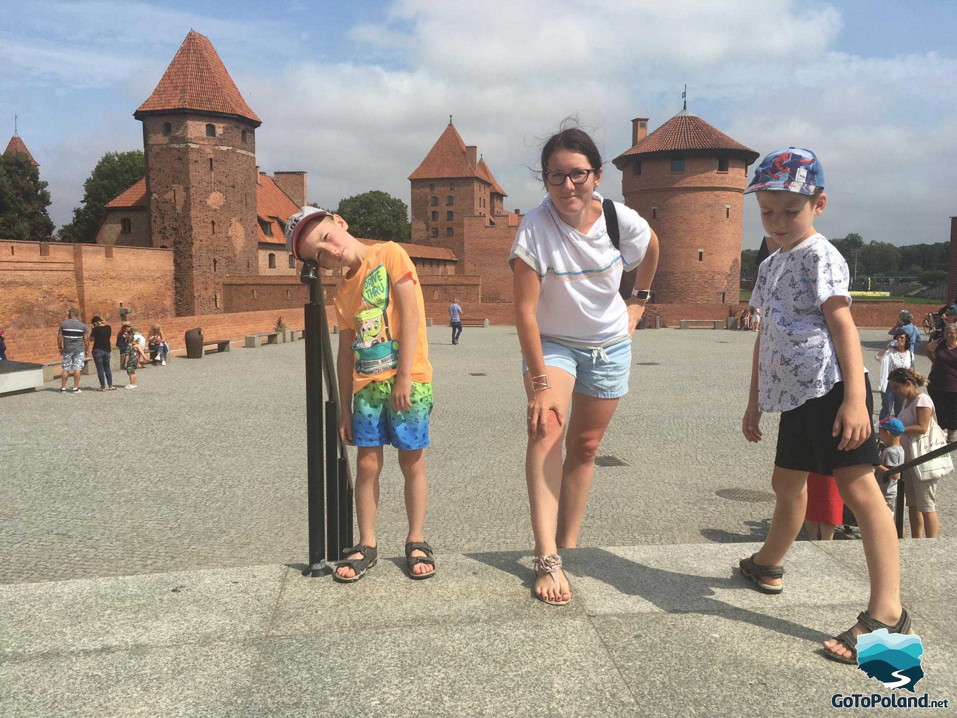 in the foreground is a woman and two boys, behind them there is a brick castle