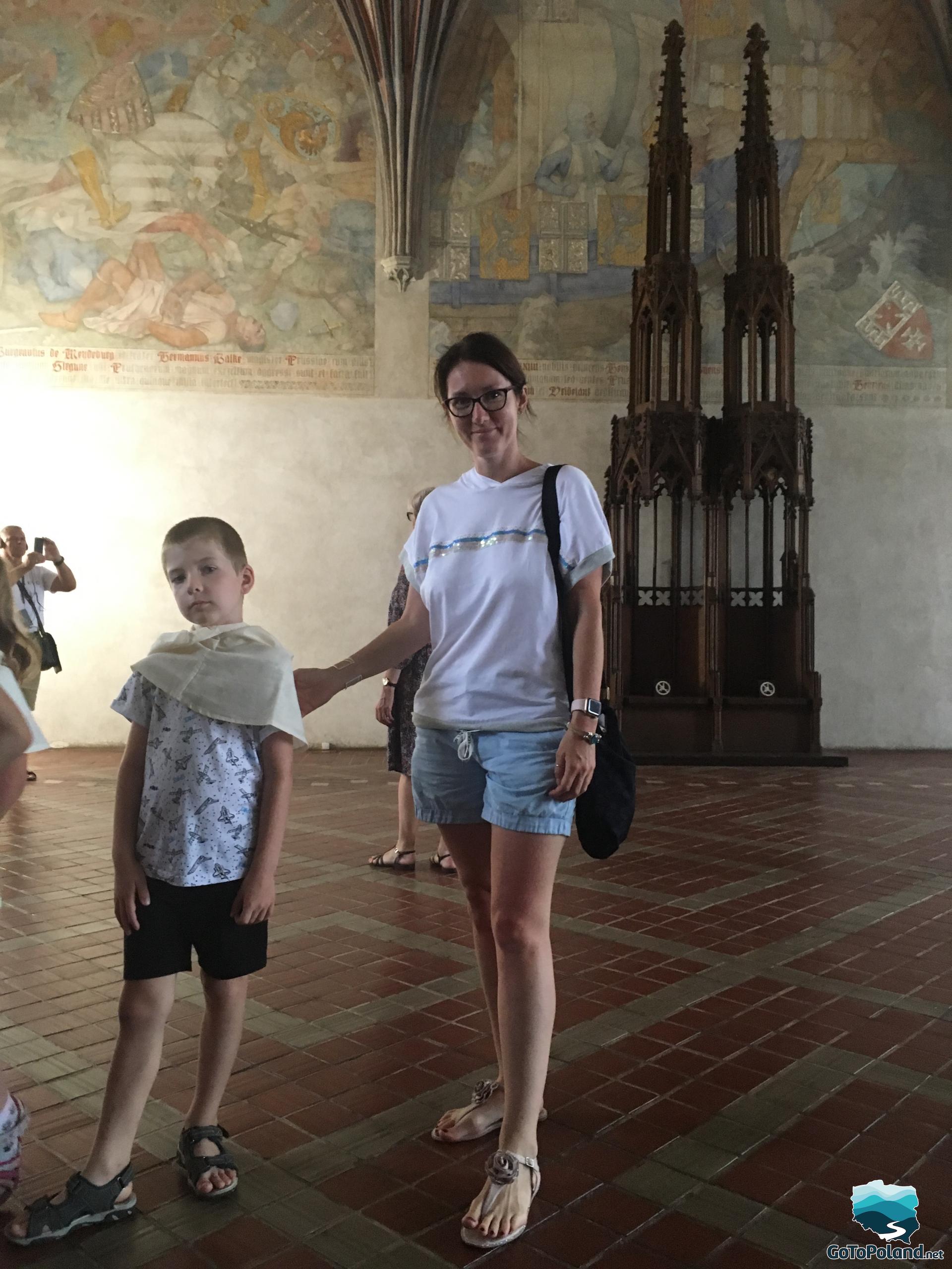 a woman and a boy standing in a castle hall, several frescoes on the walls