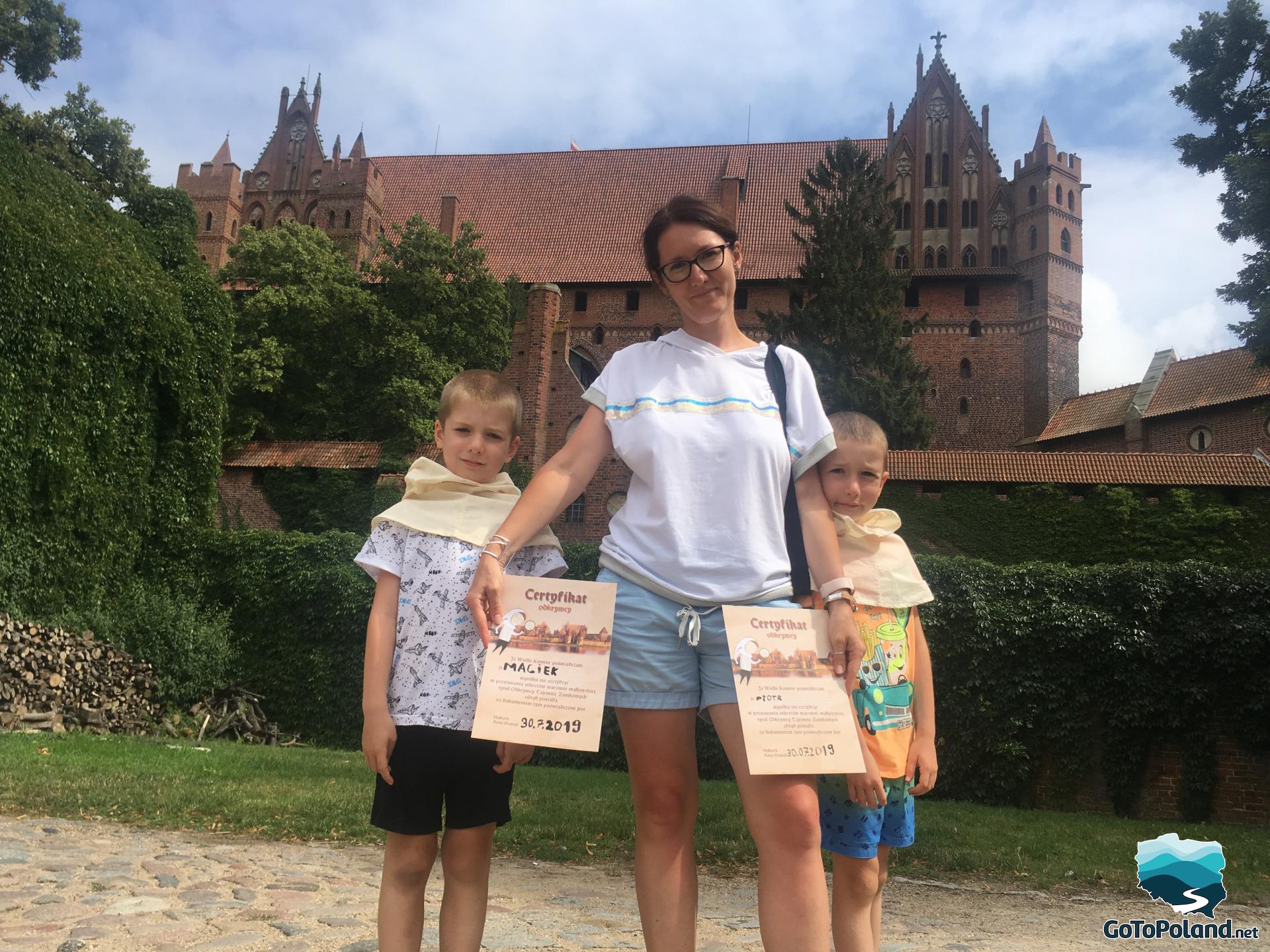 a woman and two boys are in front of a castle, a woman is holding some certificates