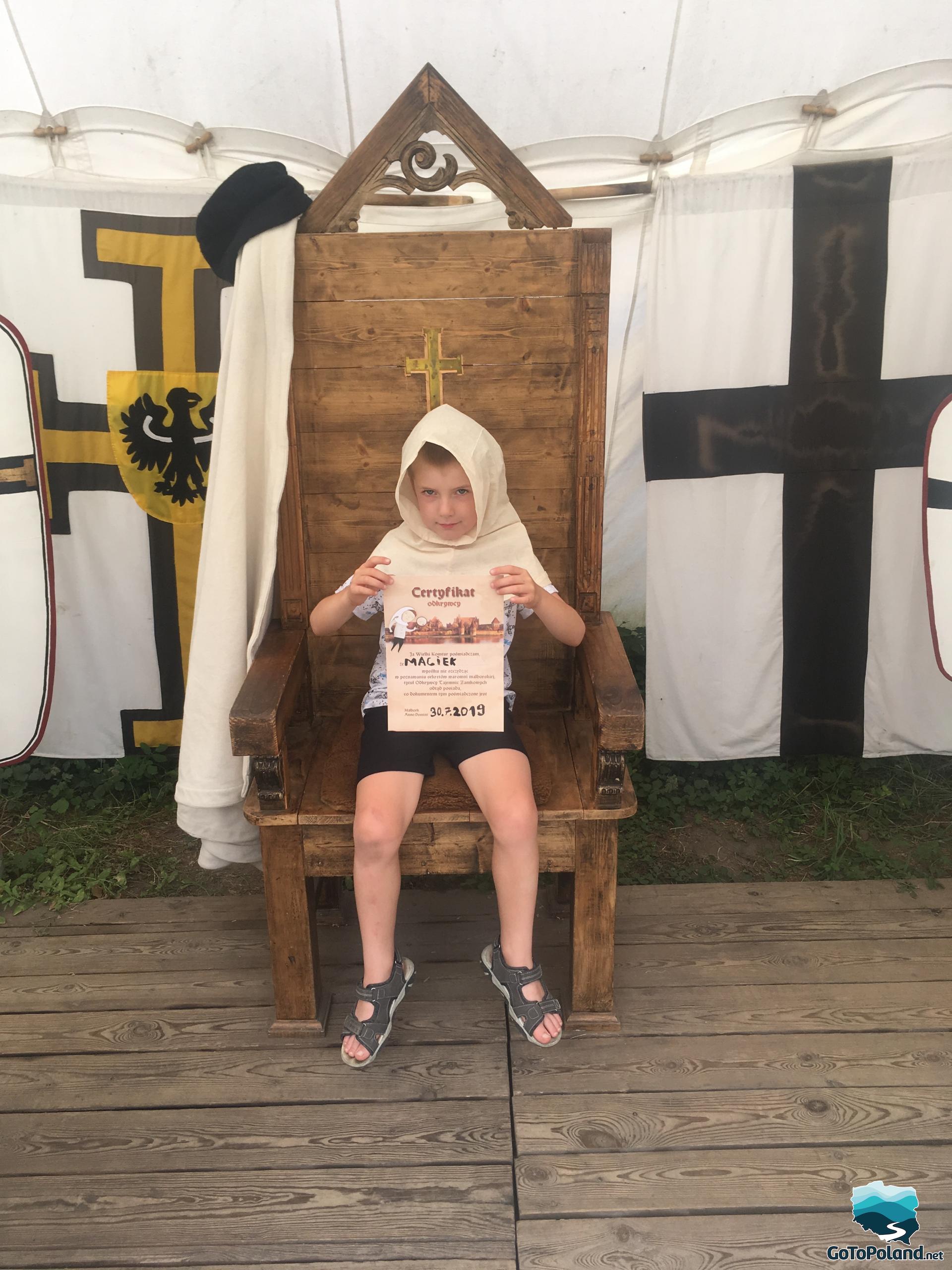 a boy is sitting on a wooden chair and holding a certificate of a discoverer
