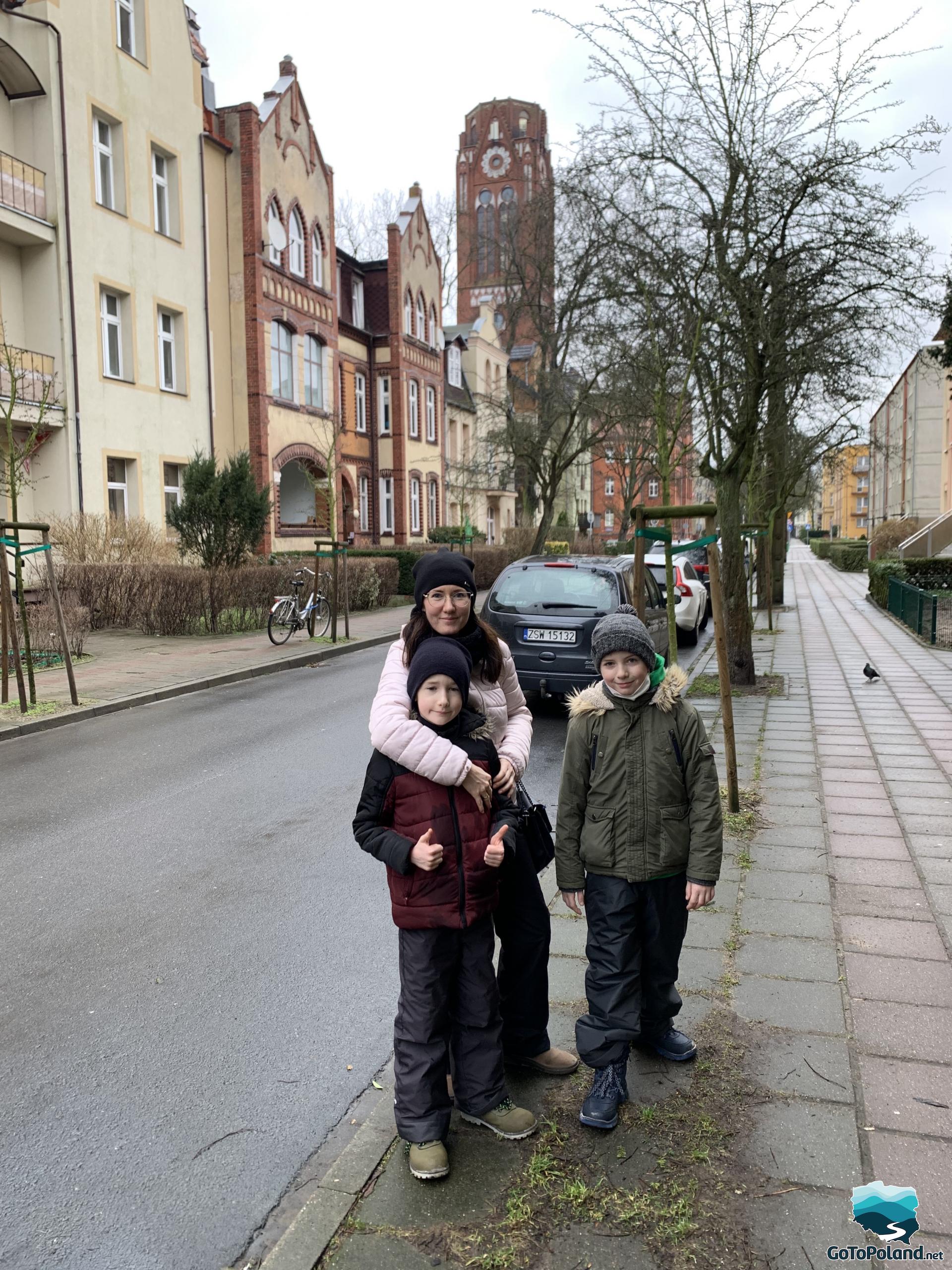 a woman and two boys are standing in the street, there are blocks of flats on both sides of the street