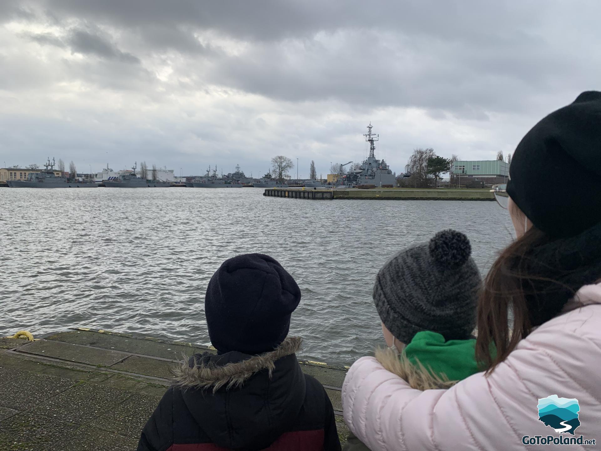 a woman and two boys are are watching the other bank of the river and navy ships
