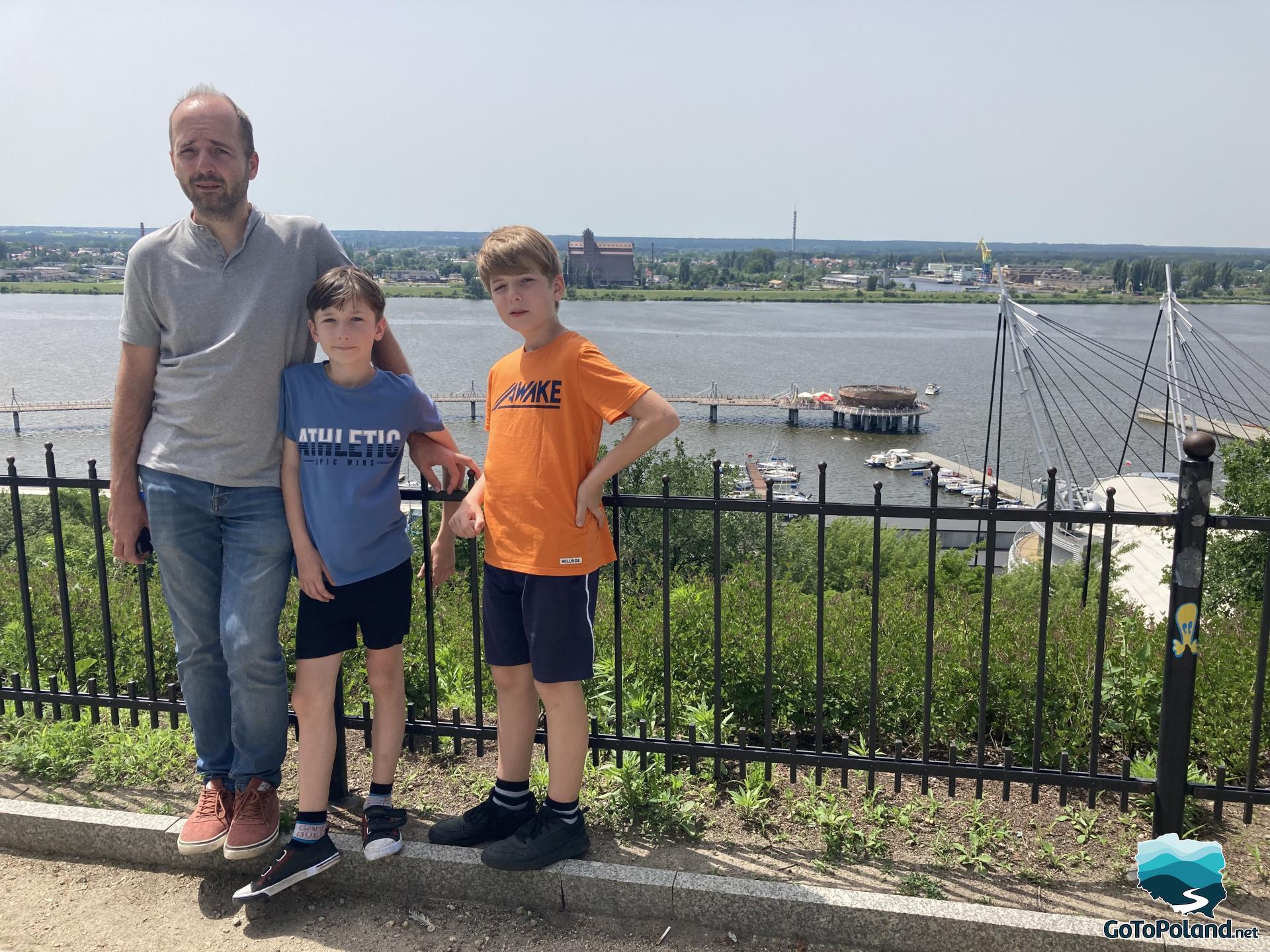 A man and two boys standing on a viewing platform. View of the river and the pier parallel to the shore in the background  