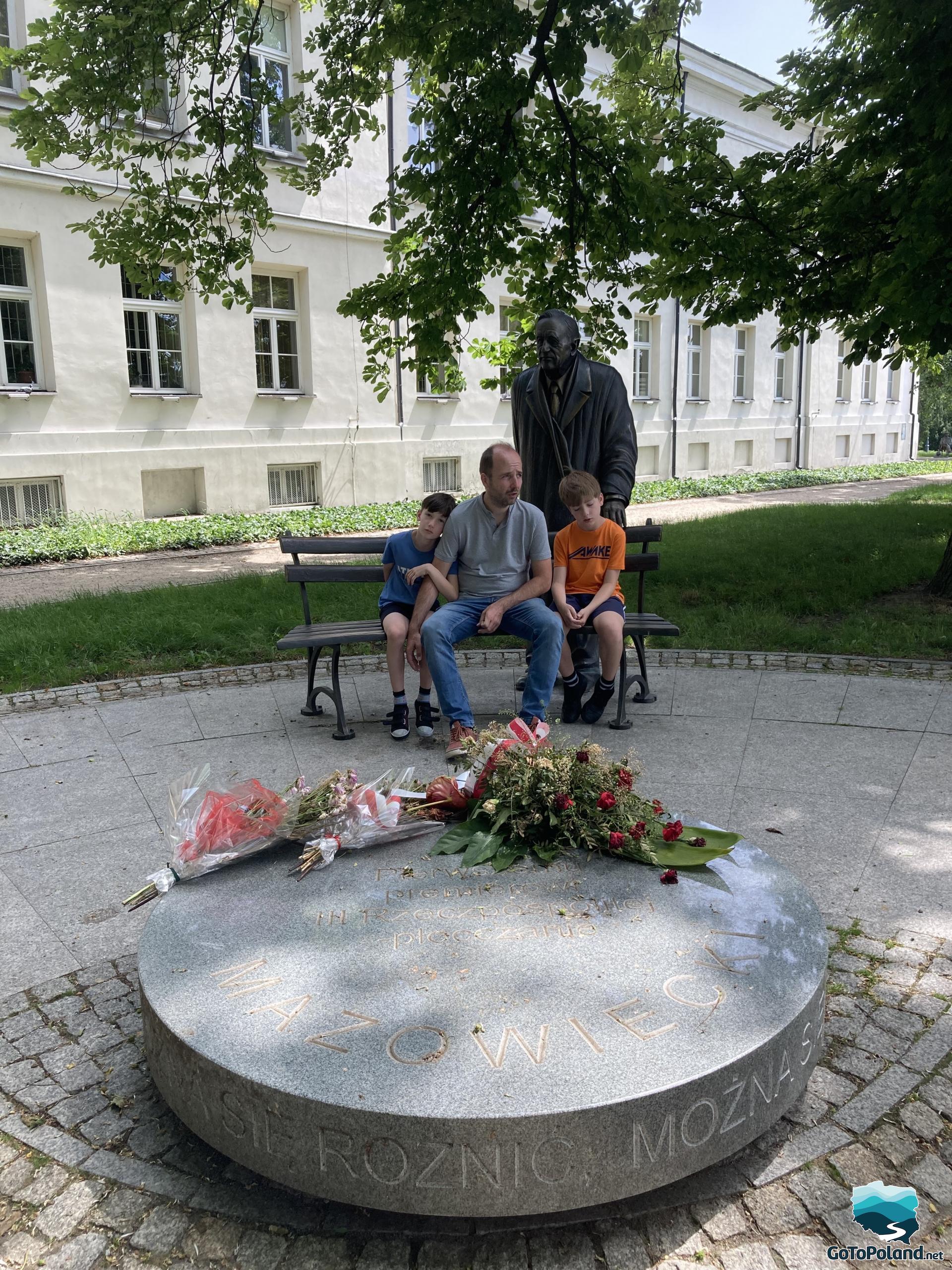 A man and two boys sitting on a bench by the monument to the first Polish prime minister after the fall of communism