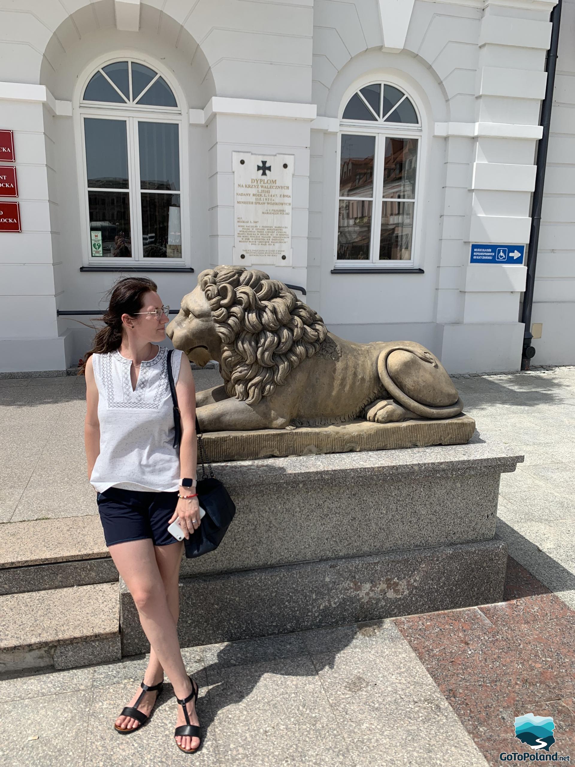 A woman standing by the lion sculpture, a lion is in front of the white building