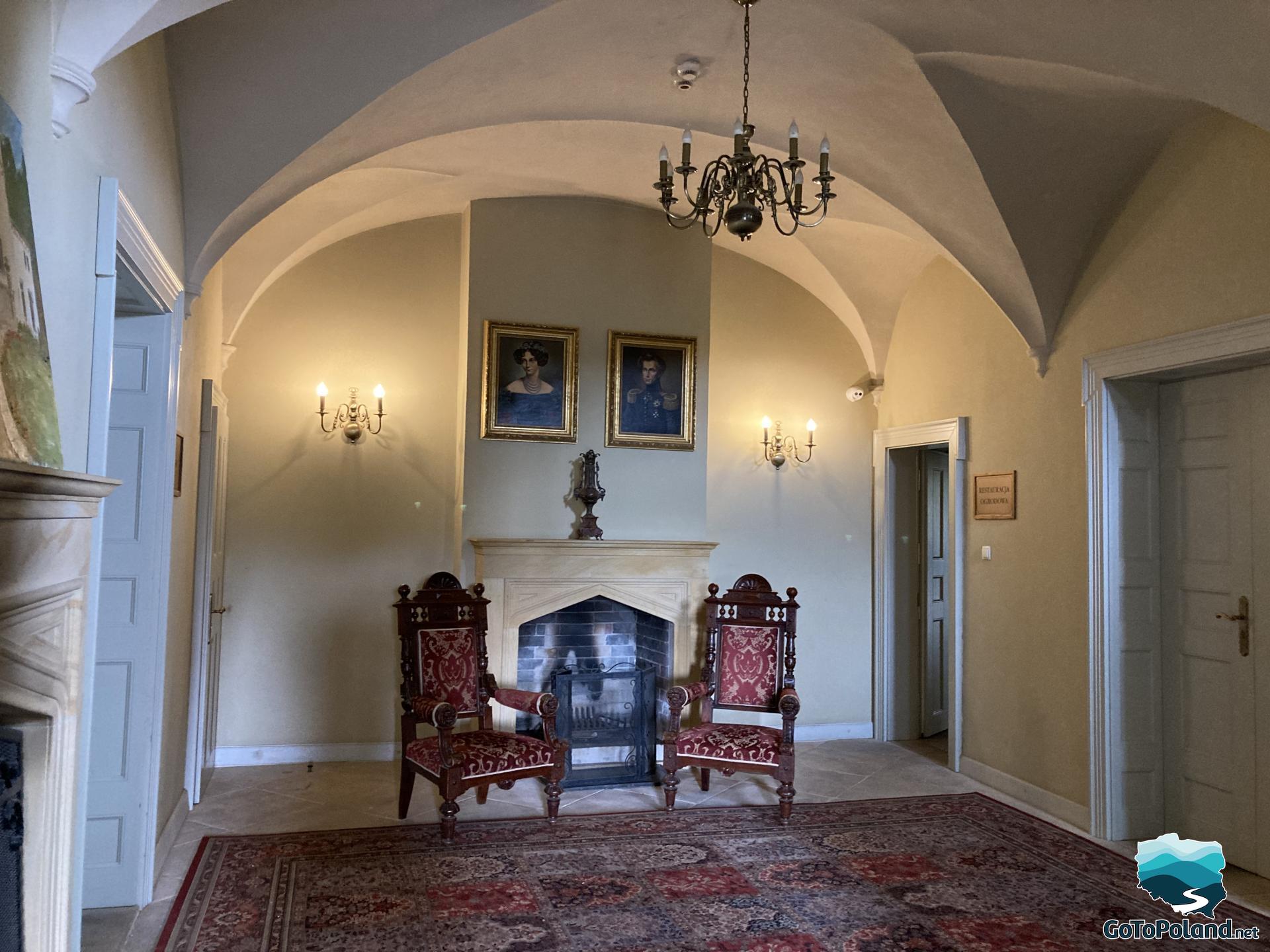 A room inside the palace, there are two armchairs by the fireplace. Two portraits hang above the fireplace on the wall