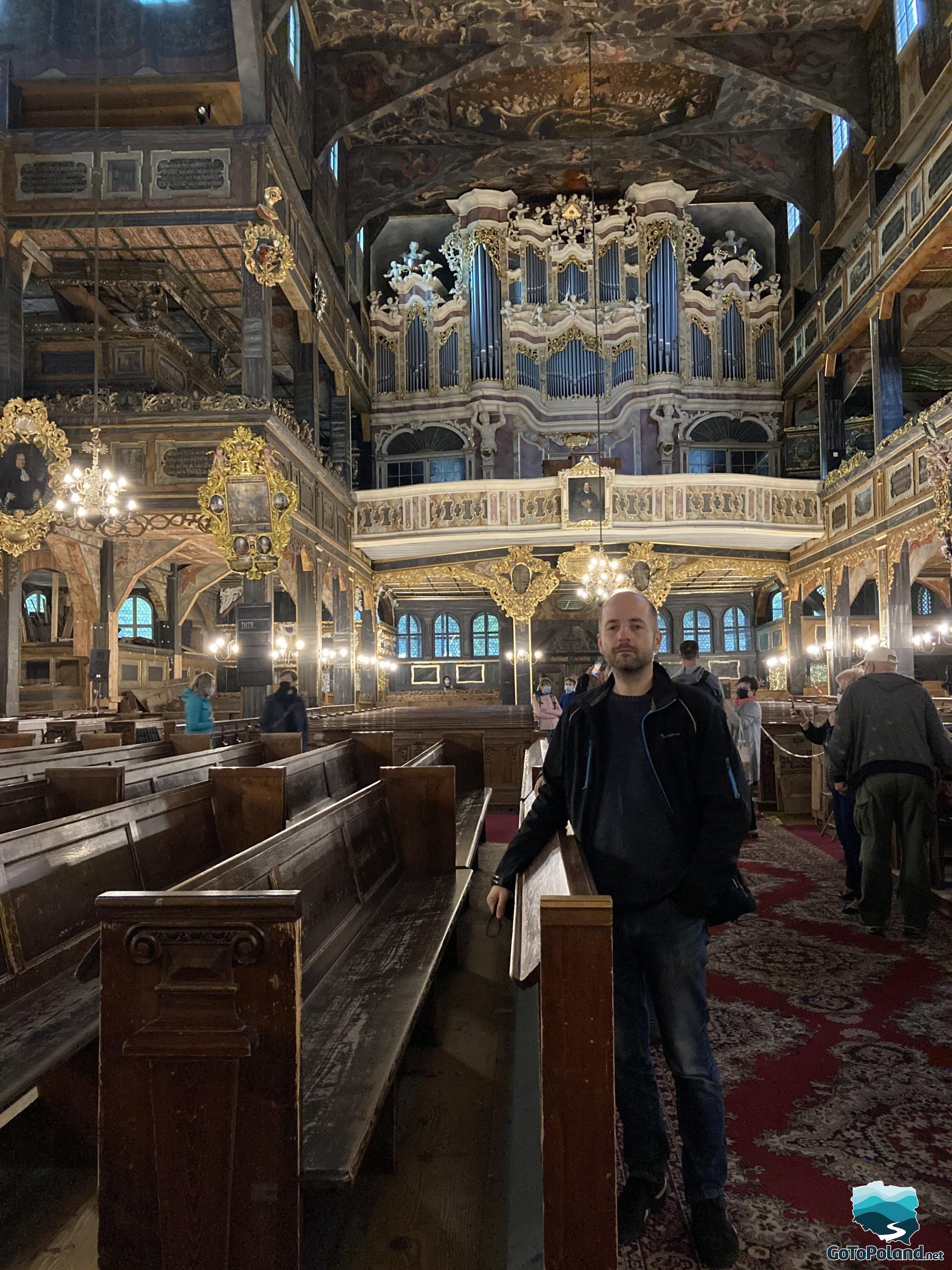 A man is standing by the wooden bench in a church, painted ceiling and the organ in the background 