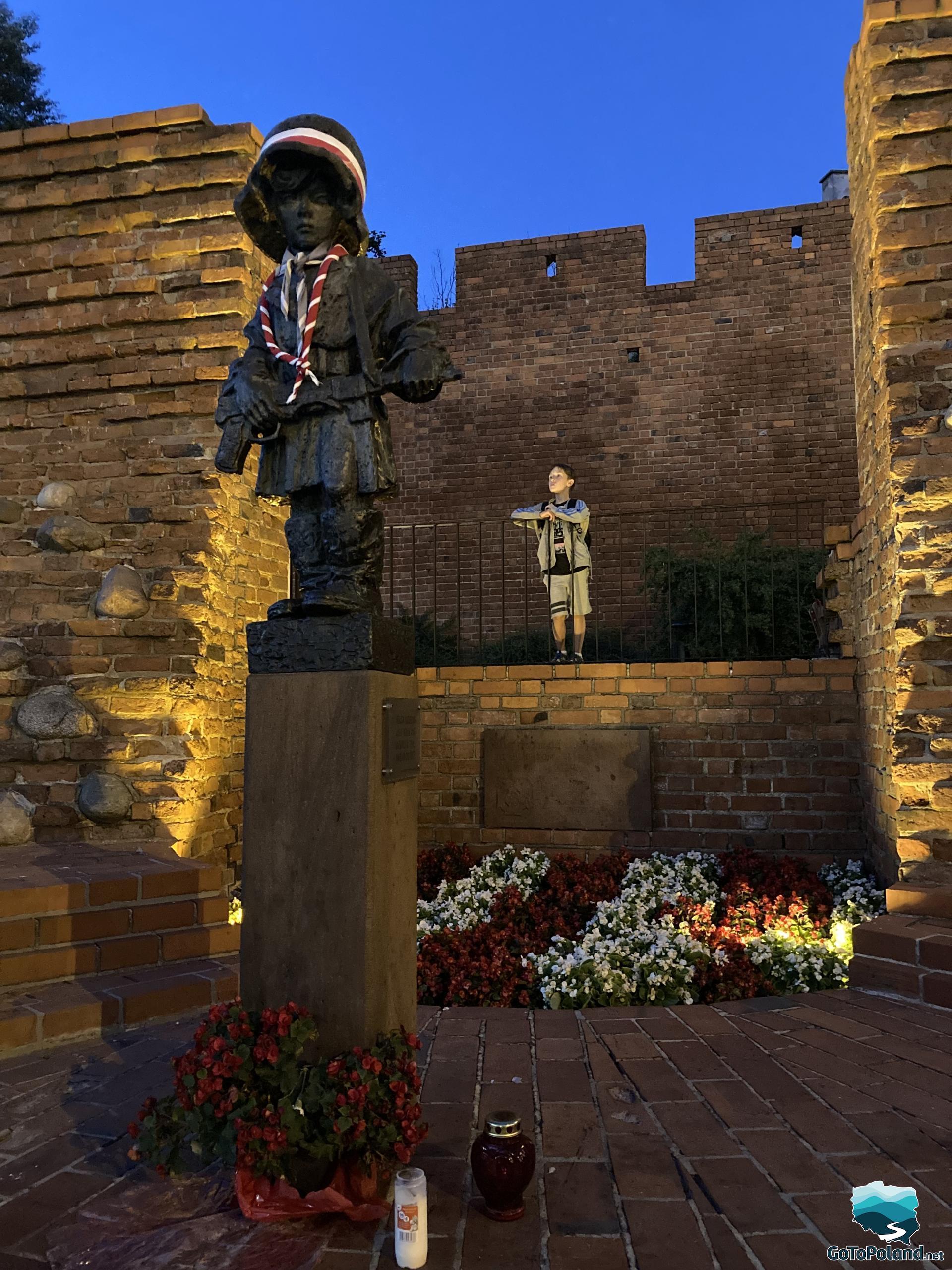 a statue of a little boy in a helmet, a little insurgent, red flowers under the statue