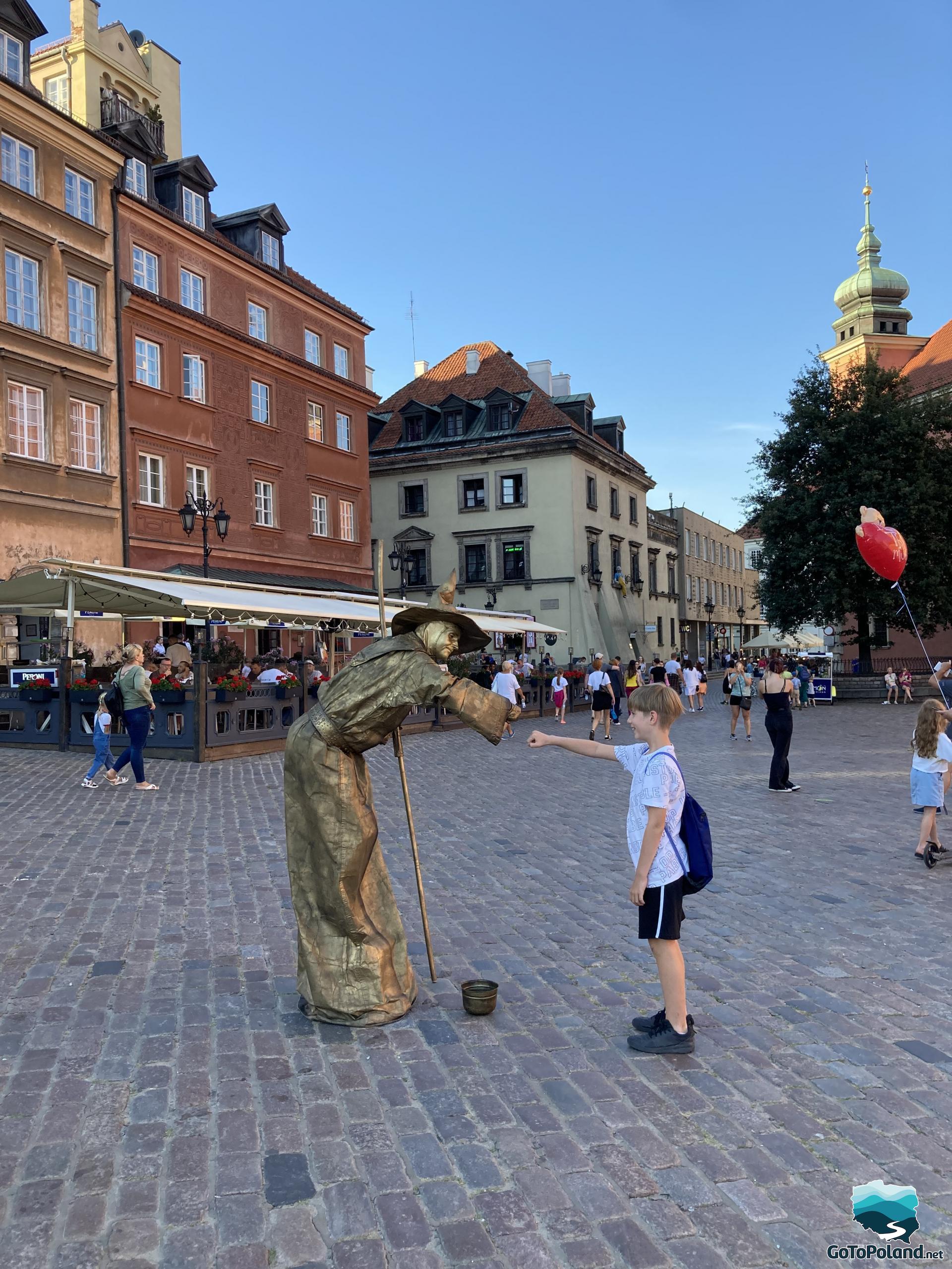 a gold-painted performer greets a boy, tenement houses in the background