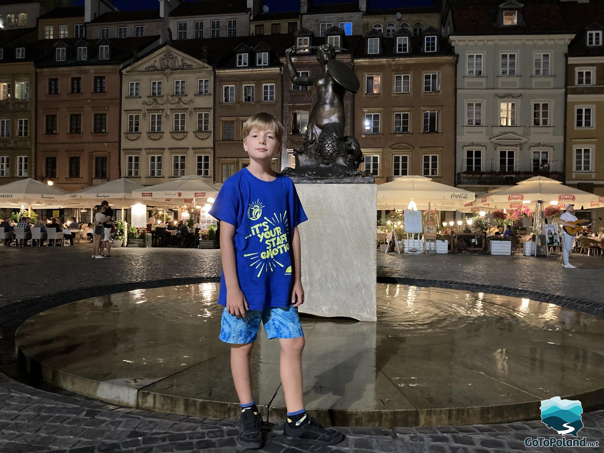 a boy is standing in front of the marmeid statue, tenement houses behind this statue