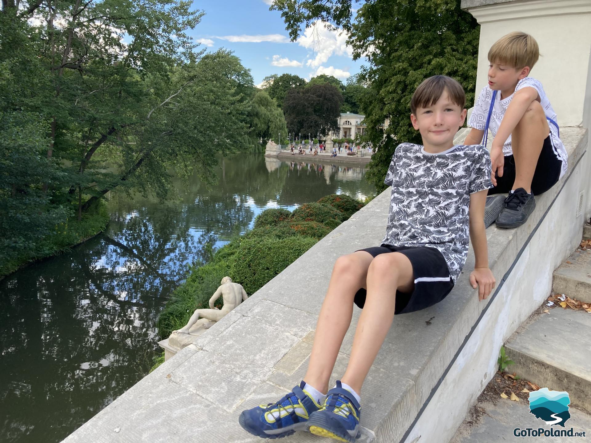 two boys sitting on the stone balustrade above a pond, a palace in the background 
