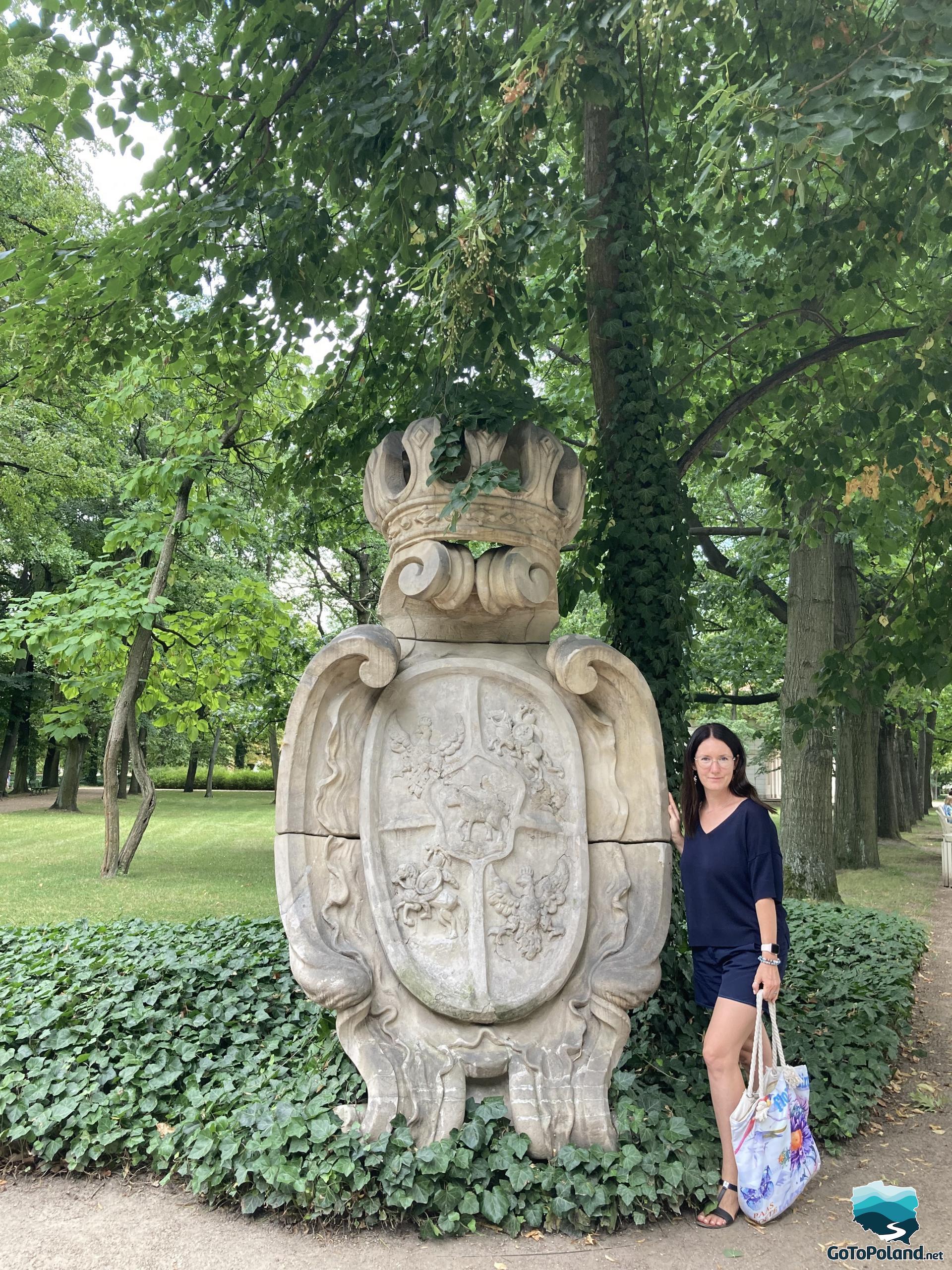 a woman stands by a stone cartouche with a coat of arms, trees in the background