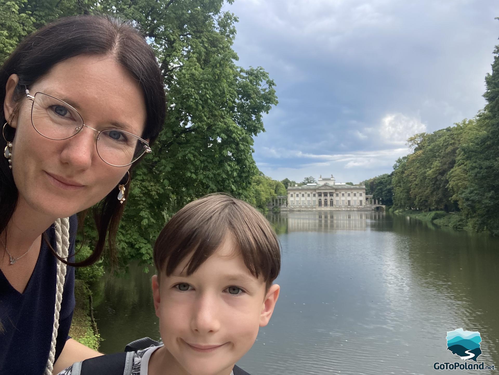 a woman and a boy are taking a selfie, behind them there is a pond an a palace
