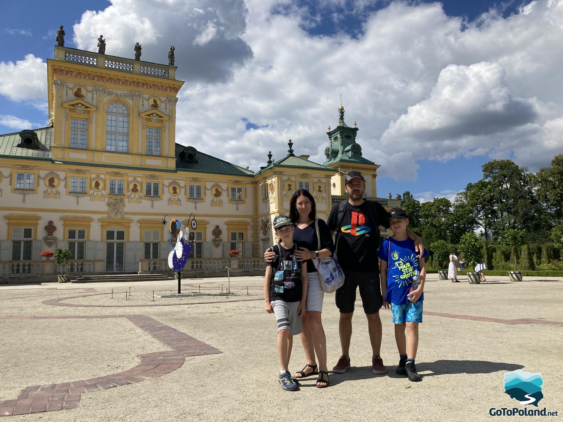 A woman, two boys and a man are standing in front of the baroque palace, the palace has yellow walls, and Roman style statues on top