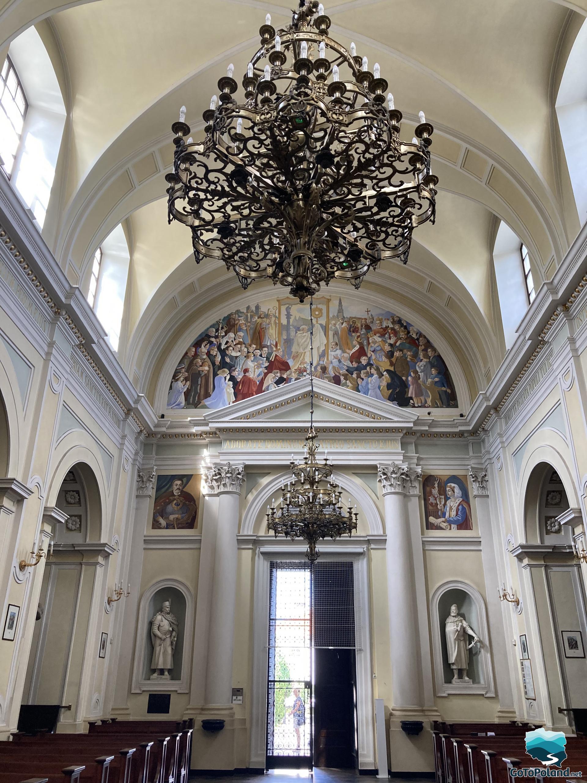 the main entrance to the church seeing from the inside, a large chandelier hangs from the ceiling, there are two statues on either side of the entrance