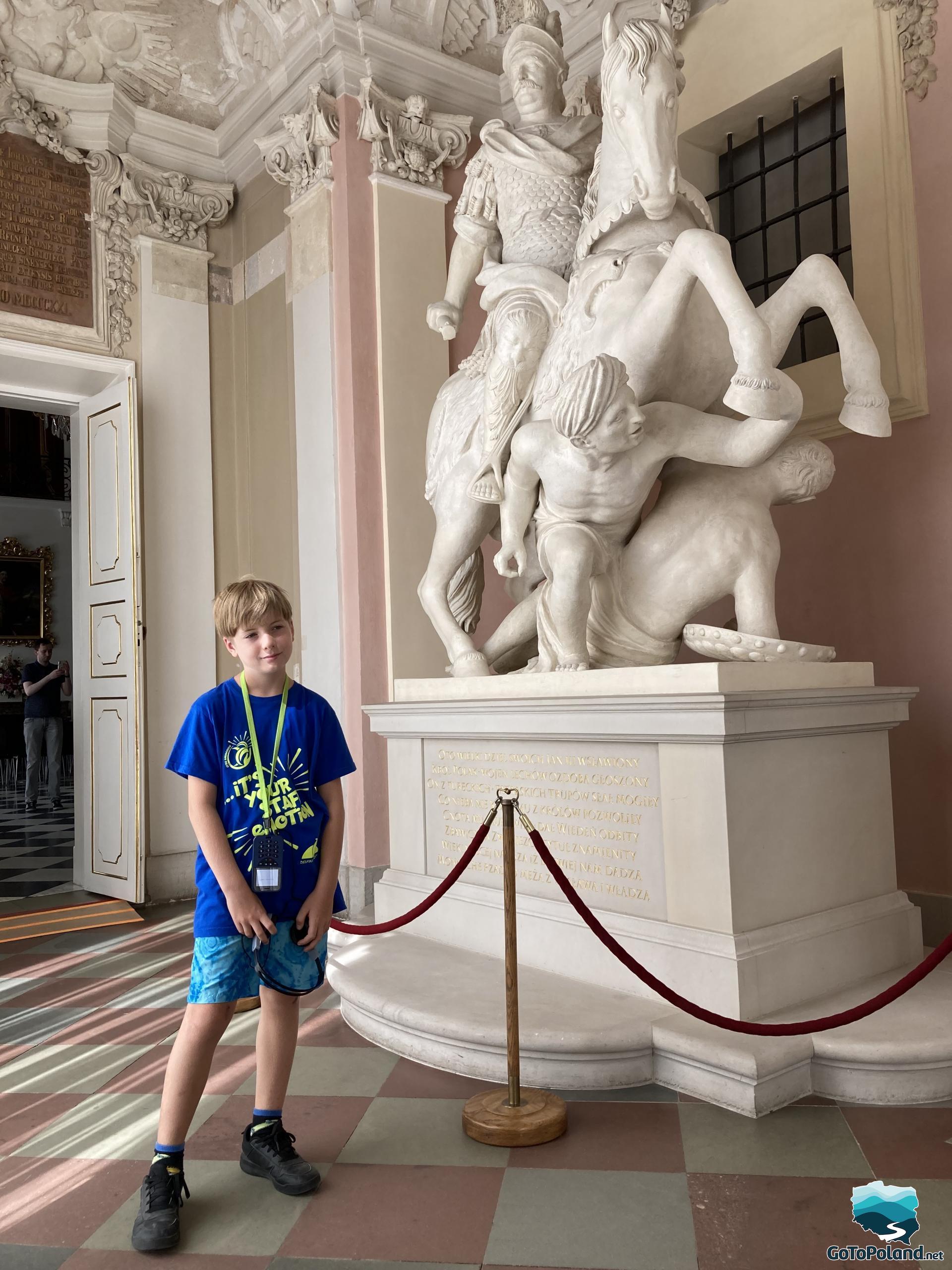 a boy is standing next to huge statue which presents a ruler on the horse. A statue is white