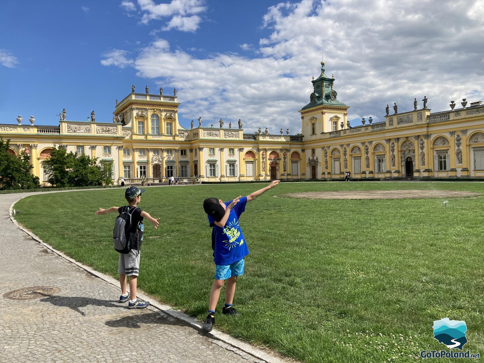 two boys are standing on an alley of cobblestones, behind them a green lawn and a large palace with a yellow facade