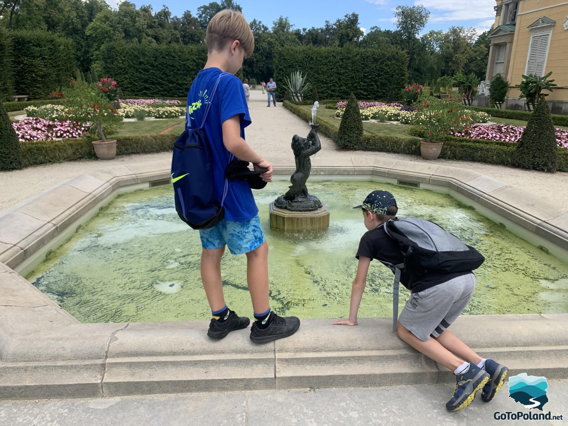 two boys are near the fountain, in the center in fountain there is a small cupid, the fountain are in the palace garden