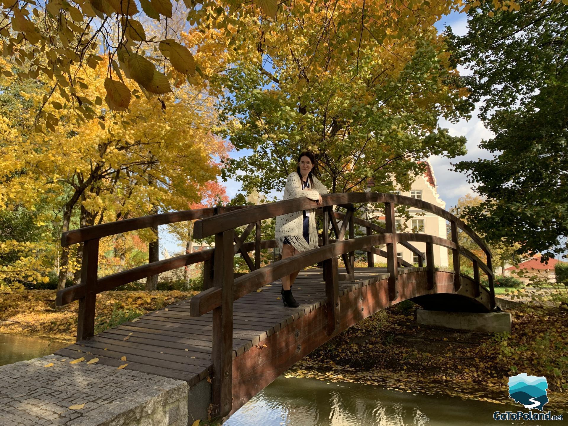 A woman standing on a small wooden bridge, trees with colourful leaves around her