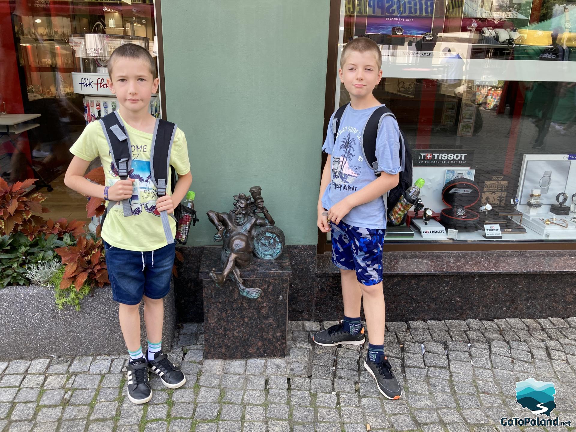 Two boys standing in from of the watch shop. They are next to a small Bacchus figurine 